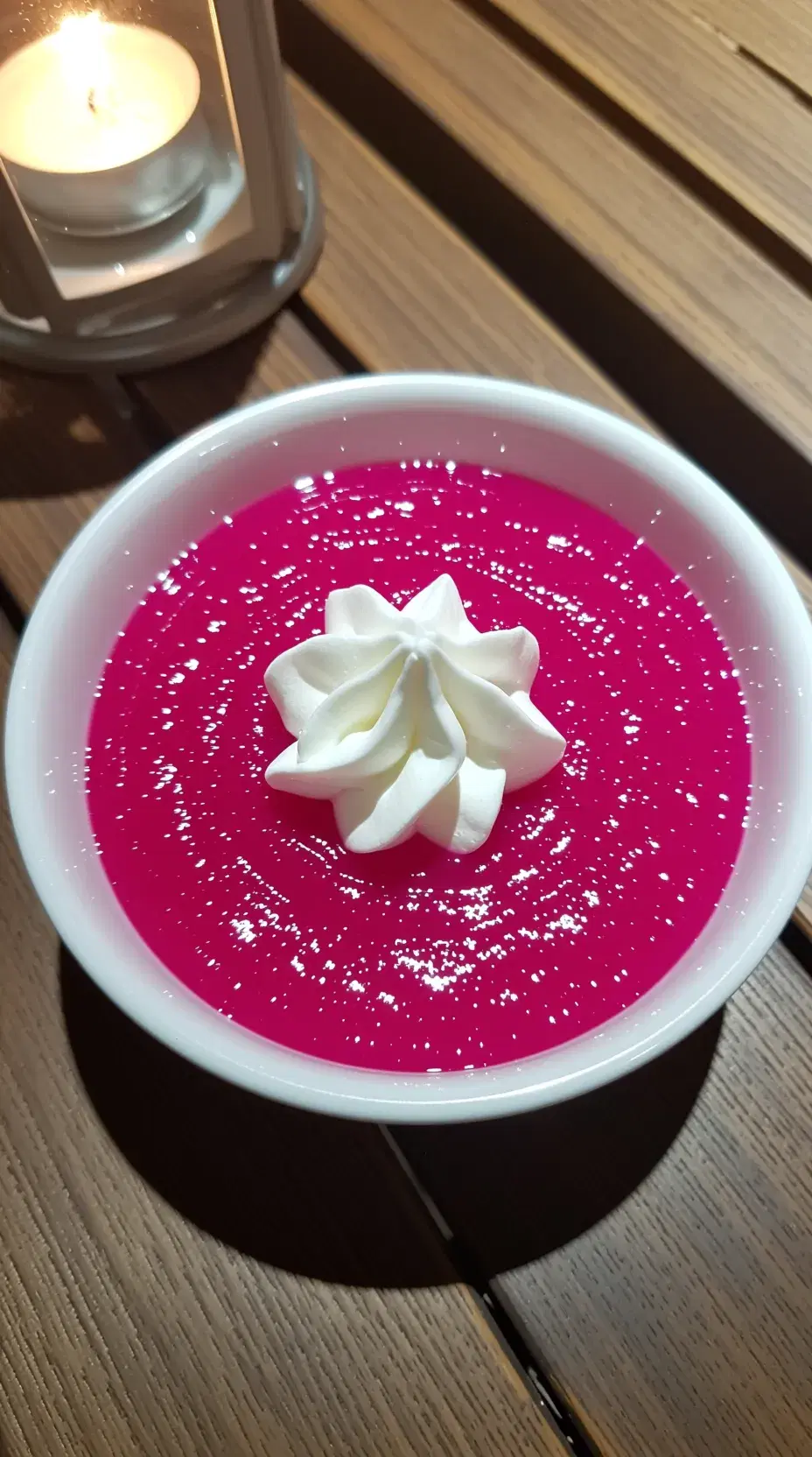 A vibrant bowl of bright pink pudding, with a dollop of whipped cream on top, set against a rustic wooden table, with a warm, inviting glow from a nearby candle.